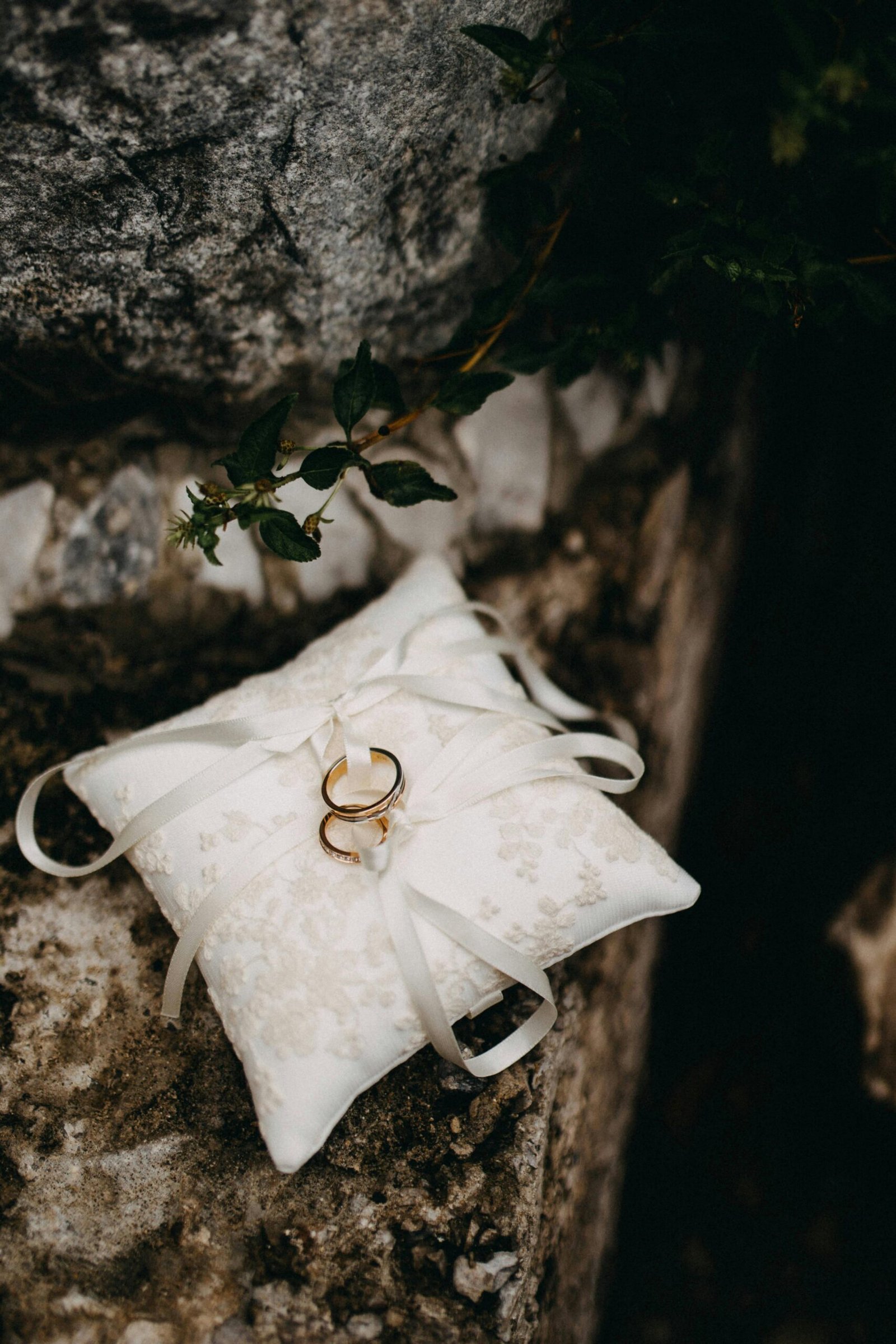 Close-up of wedding rings on a silky white cushion outdoors, captured in soft light.
