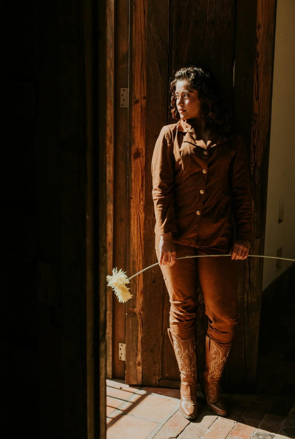 A woman in brown clothing stands indoors holding a long-stemmed flower, lit by sunlight.